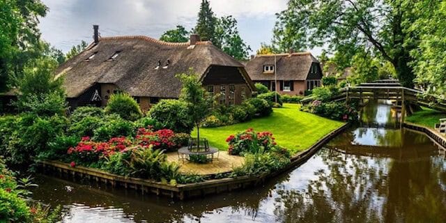 A beautiful canal-side cottage in Giethoorn with a thatched roof, surrounded by a lush garden, flowers, and a wooden footbridge over the water