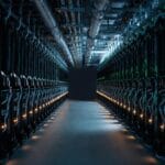 A wide-angle interior view of a massive AI Infrastructure data centre aisle lined with rows of dark, industrial server racks illuminated by rhythmic status lights, highlighting the scale and complexity of AI hardware